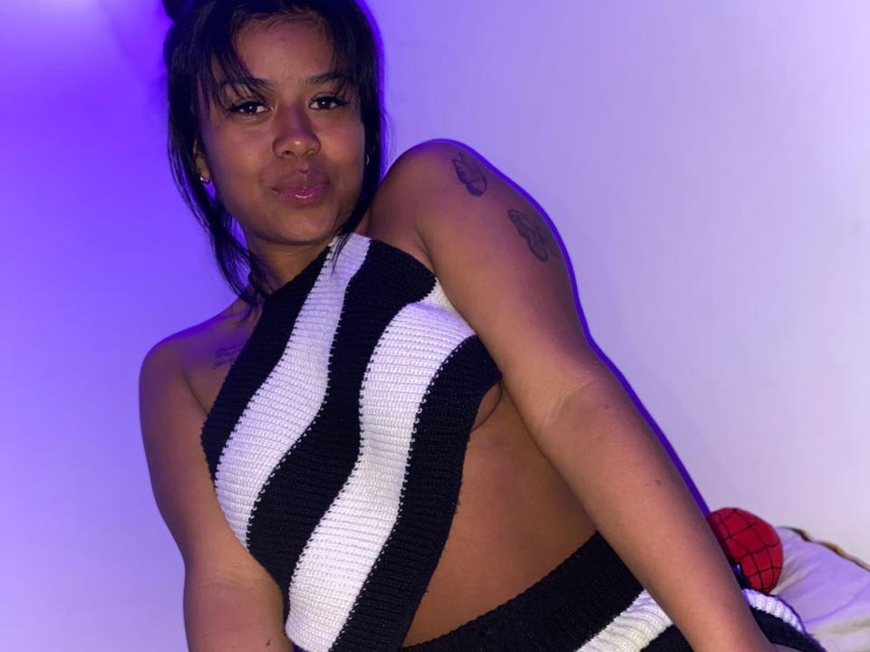 EmmaWater with brown skin and black hair, wearing a black and white striped halter top, showing her small tits and tattoo on her right arm, pouting her lips.