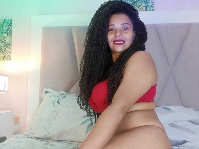 KenyaKarath with curly black hair, red bra, and no panties, smiling on a bed, showing her ass and thick thighs, in a bedroom with a tufted headboard.