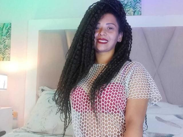 KenyaKarath with long, black, curly hair wearing a white, hexagonal-patterned top, exposing pink, round boobs, smiling, standing in a bedroom, with a grey upholstered headboard.