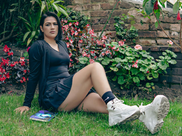 ElizabethDupont with black dress, white sneakers, and black socks, sitting on grass, legs crossed, showing her ass, surrounded by red flowers, holding a colorful book.