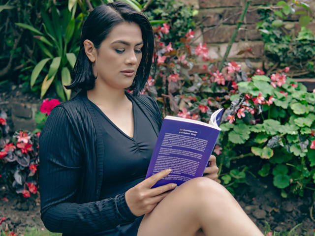 ElizabethDupont with black hair, black dress, and cardigan, reading a purple book, sitting with crossed legs, showing her tits, in a green, plant-filled garden.