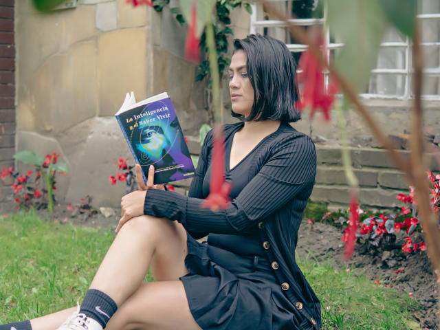 Elizabeth Dupont mit schwartem Bob, schwarzem Pullover, schwarzem Kleid und Socken, sitzend auf dem Gras, ein Buch lesend, Beine gekreuzt, wobei ihr Hintern sichtbar ist, umgeben von roten Blumen, ernster Gesichtsausdruck..