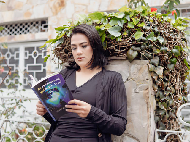 ElizabethDupont with dark hair, black ribbed shirt, and black pants, reading a book with a blue cover, standing in front of a stone wall with green vines.