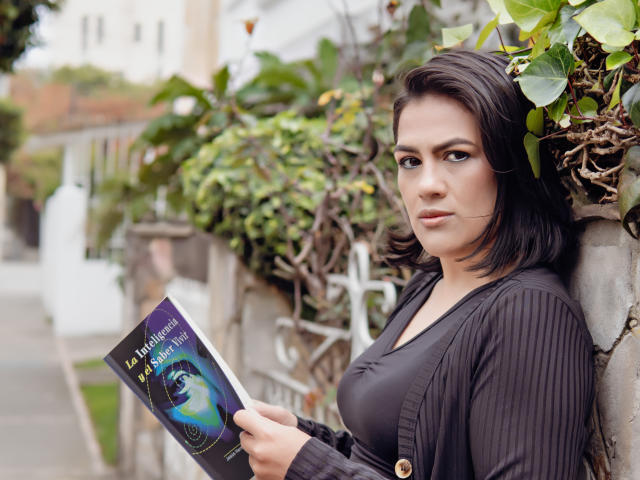 ElizabethDupont with black hair, looking at a book, holding it in her right hand, wearing a black top, leaning against a wall with green vines, her tits visible.
