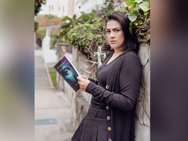 ElizabethDupont with dark hair, holding a book, wearing a black ribbed cardigan and dress, standing against a wall, green leaves in background, looking slightly annoyed, showing her tits.