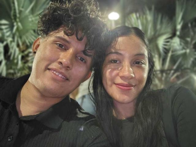 LoveAndSin, com cabelo preto encaracolado e uma camisa preta, sorrindo ao lado de LoveAndSin, com cabelo preto comprido e uma camisa preta, ambos parecendo fofos e felizes..