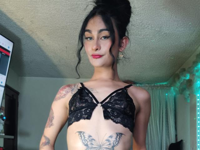 KamilaBabe with black lace bra, tattoos on arms and chest, butterfly tattoo below tits, black hair in messy bun, red lipstick, looking down, small nose ring, in room.