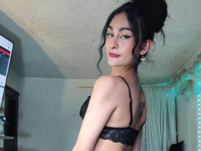 KamilaBabe with black lace bra, looking back, red lipstick, wavy hair up, small star earring, tattoo on upper back, slight smile, medium tits, light skin, bedroom.