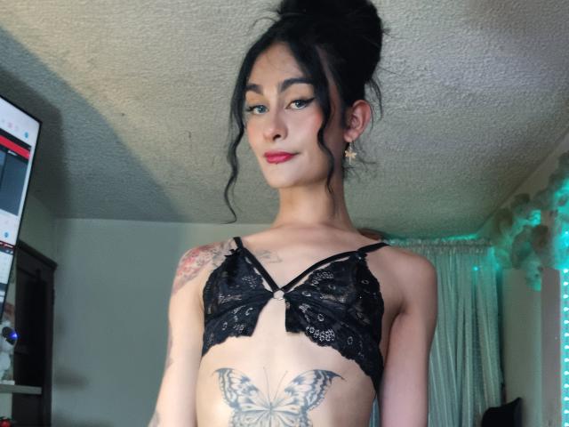 KamilaBabe with black lace bra, tattoos on shoulder and butterfly on belly, black hair in messy bun, red lipstick, green eyes, small earrings, standing in dimly lit room.