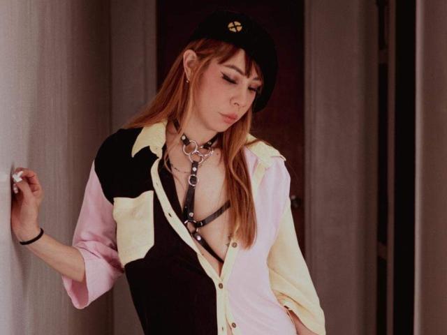 AriiannaHot with long red hair, wearing a black cap, pink and black button-up shirt, black harness, and choker, leaning against a wall, eyes closed, looking sexy.