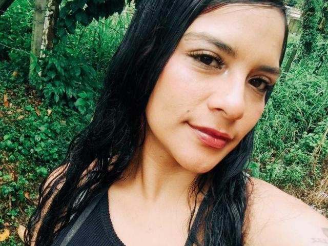 Mia Bellucci tem cabelo preto ondulado, olhos castanhos e batom vermelho. Ela está vestindo uma camiseta preta sem mangas, que mostra seus pequenos seios, e tem um leve sorriso no rosto..