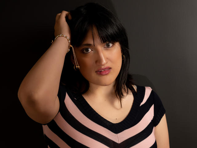 CarolineHarpper with black hair, pink and black striped shirt, hand in hair, brown eyes, pink lips, gold earrings, gold bracelet, standing against dark background, looking at camera.