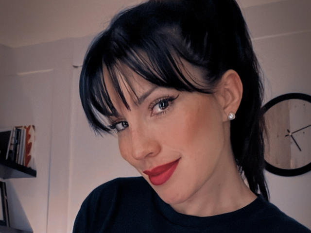 Liliysweet with black hair, bangs, and red lipstick, wearing a black shirt, smiling, blue eyes, diamond earrings, and fair skin. Clock and bookshelf in the background.