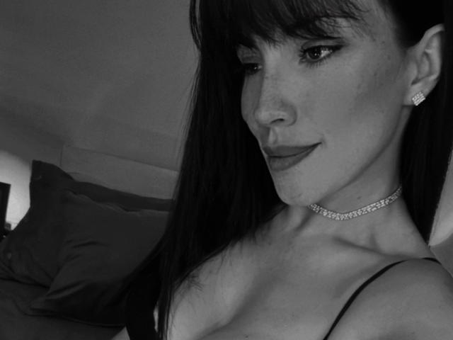 Liliysweet with dark hair, bangs, and earrings, wearing a choker, smiling slightly, showing her tits, in a black-and-white selfie.