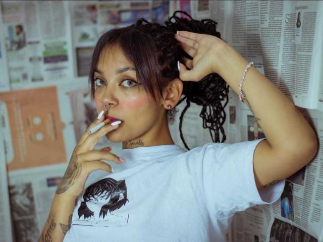DulceTammara69 with dark curly hair, nose ring, and tattooed neck, wearing a white tee, biting a cigarette, hand in hair, bracelet, and tattoos, looking at camera.