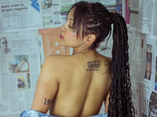 Topless DulceTammara69 with long braided ponytail, crown tattoo on upper back, "L" tattoo on left arm, pink lipstick, looking over shoulder, newspaper-covered wall in background.