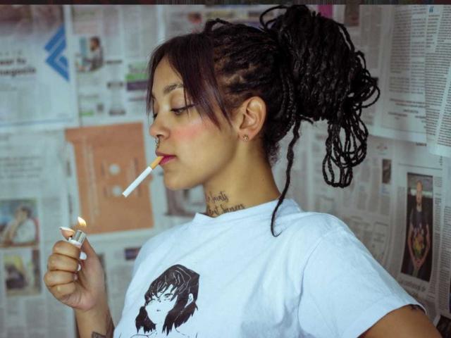 DulceTammara69 with dark braids in a messy ponytail, smoking a cigarette, wearing a white tee with a black girl graphic, nose ring, and tattooed neck.