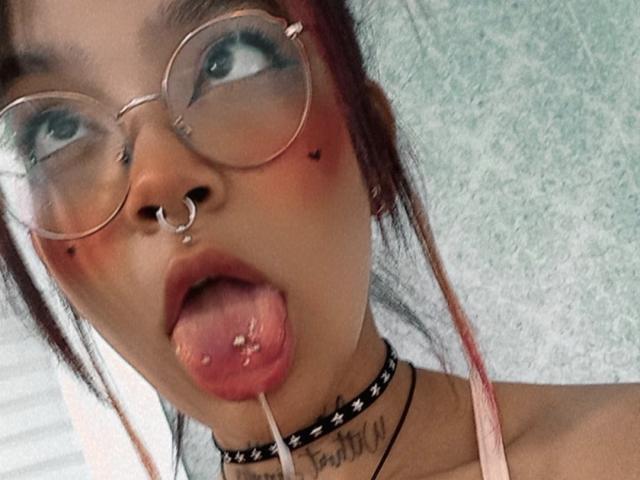 DulceTammara69 has pinkish-red hair, glasses, nose ring, septum ring, and black choker. Tongue out, with a straw in mouth, and a tattoo on her neck.