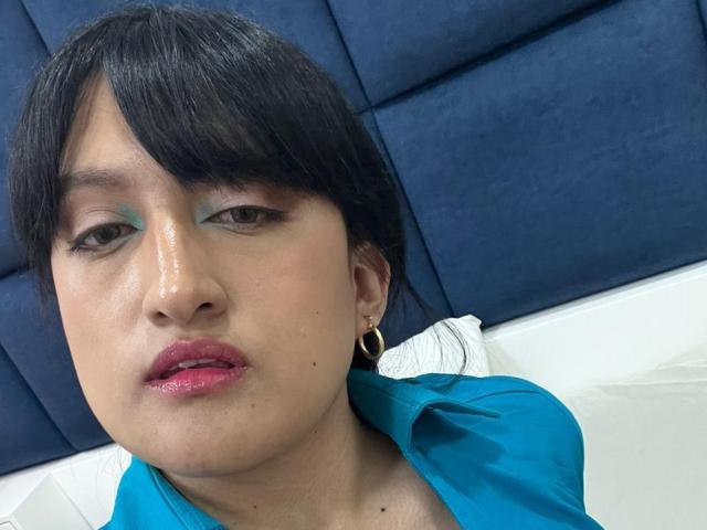 CarolineHarpper with shiny skin, black hair, green eyeshadow, pink lips, gold hoop earrings, blue shirt, looking at camera, slightly sweaty, on white pillow, blue headboard in background.