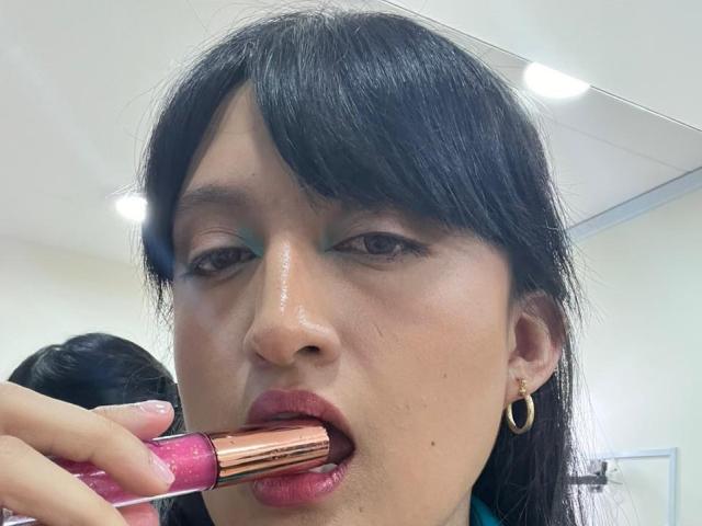 CarolineHarpper's face, with black hair and gold hoop earrings, applying pink lipstick to slightly parted red lips. She has green eyeshadow and shiny skin. Her hand holds the lipstick.
