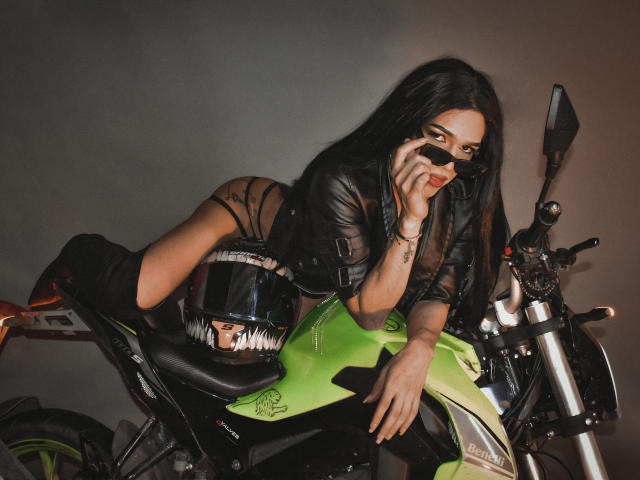 StefyFerrer with long black hair, leather jacket, and black boots, leaning on a green motorcycle, holding a phone to her ear, wearing a black helmet, looking sexy.