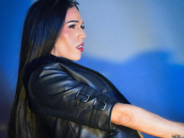 StefyFerrer with long black hair, wearing a black leather jacket, side profile, looking to the left, red lipstick, hoop earrings, blue-lit background, sexy, confident, attractive.
