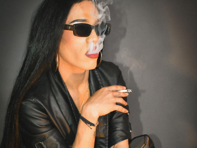 StefyFerrer with long black hair, black leather jacket, black sunglasses, smoking a cigarette, white smoke, red lipstick, gold hoop earrings, black bracelet, holding cigarette in right hand.
