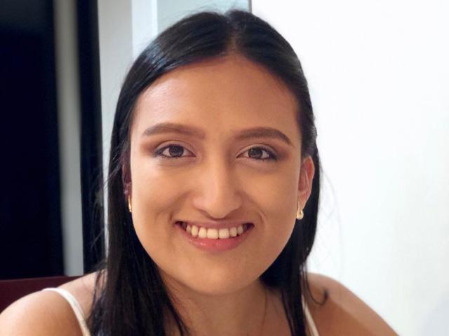 CarolineHarpper with straight black hair, brown eyes, smiling, wearing a white tank top, showing her tits slightly with her arms down. She's wearing small pearl earrings.