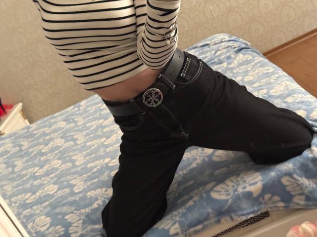 Siyana with striped shirt, black jeans, and belt, kneeling on a blue-patterned bed. Visible belly button, black belt with pentagram buckle. Room has wooden floor, wallpaper.
