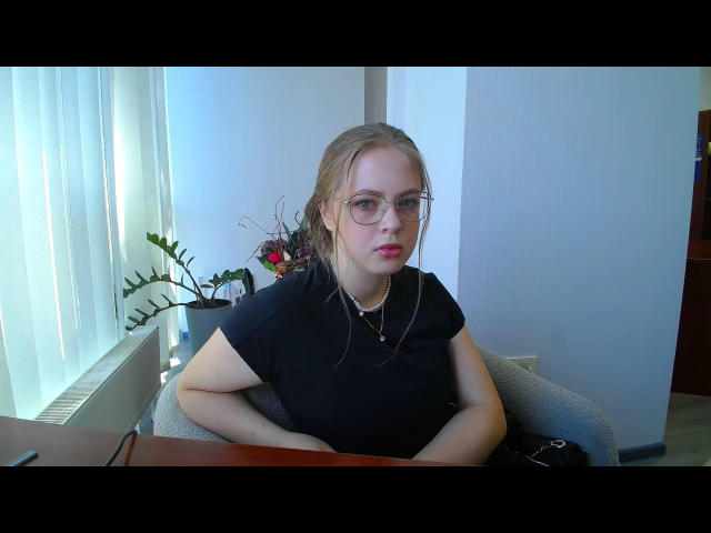 Ministiya with glasses, red lipstick, black shirt, pregnant belly, arms crossed, sitting in chair, looking at camera, small flower vase in background, white wall, vertical blinds.