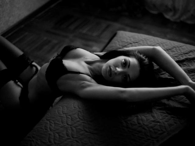 LolaBlaze, con un aspecto sensual, está acostada en una cama, con los brazos por encima de la cabeza, vistiendo lencería negra con tirantes, mostrando sus pechos y exhibiendo su cuerpo sexy, delgado y atractivo en una fotografía en blanco y negro..