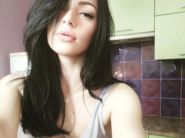 LolaBlaze, con cabello negro largo, labios rosados y finos, y un pequeño busto, vestida con una camiseta gris, mirando ligeramente hacia un lado con una sonrisa sutil y sensual..