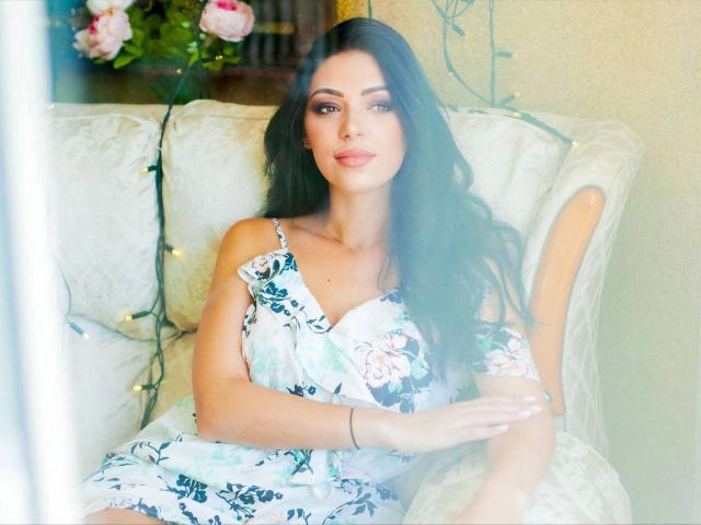 LolaBlaze, con el pelo largo y negro, vestida con un vestido floral, sentada en un sofá blanco, mirando a la cámara con una sonrisa sutil, con luces de hadas en el fondo..