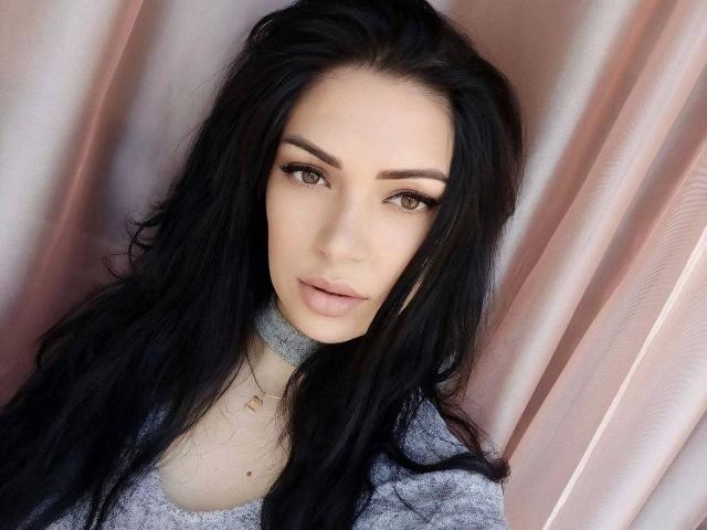 LolaBlaze-hot has long black hair, brown eyes, and full pink lips. She's wearing a gray sweater with a subtle pattern, and a delicate gold necklace.