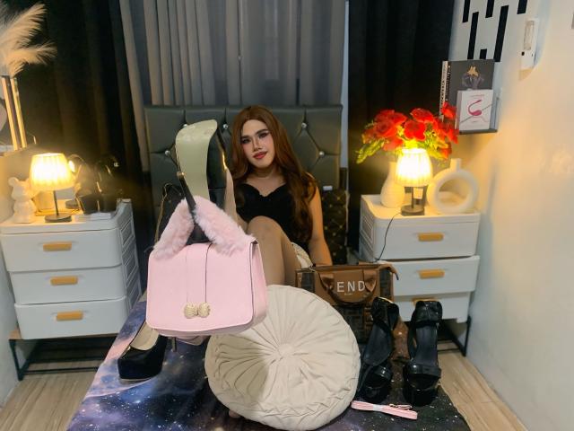 YeshaSebastian with long brown hair, black dress, and red lipstick, sitting on a bed, legs up, pink purse with fluffy ears, white cushion, black boots, and red flowers.