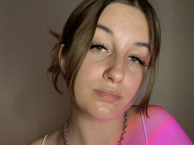 RonixHofma a des lèvres gonflées, rosées, des yeux verts avec de longs cils, et un léger sourire. Ses cheveux bruns sont attachés en un chignon désordonné. Elle porte un haut blanc avec un collier en chaîne..