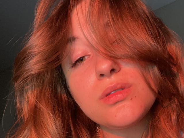 Le visage de RonixHofma, avec ses cheveux roux en désordre qui couvrent un œil, ses lèvres rosées légèrement entrouvertes, ses petites taches de rousseur et son sourire discret. Elle a un teint clair et un air adorable..