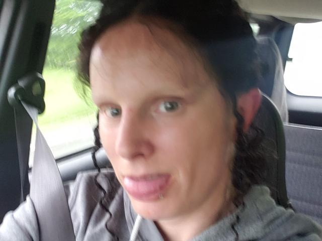 KaszyMarie has a small lip piercing on her lower left lip, with slightly parted pink lips and a small amount of facial hair. She's in a car, looking forward.