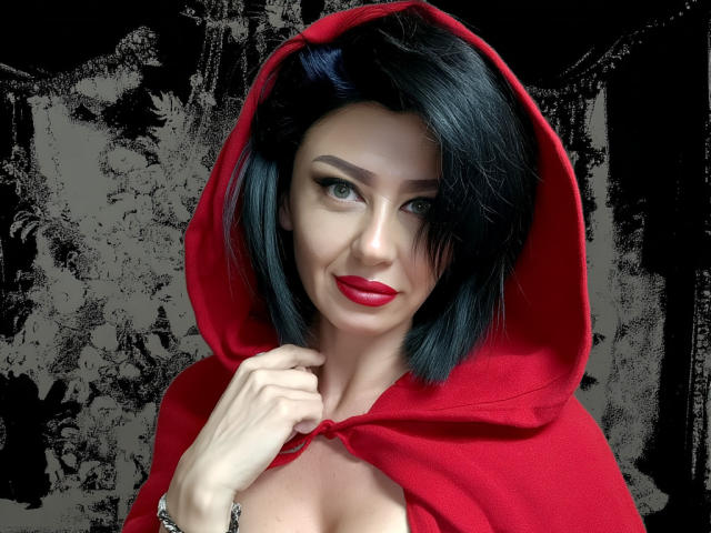 Taissa69 with black bob, red hood, and red lipstick, holding her hood, showing cleavage, green eyes, and a subtle smile, wearing a red cape.