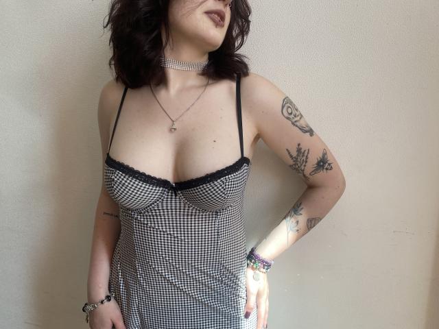 Khori with pale skin, dark wavy hair, wearing a black and white checkered dress with black lace trim, showing tits, tattoos on arms, necklace, bracelet, and choker.