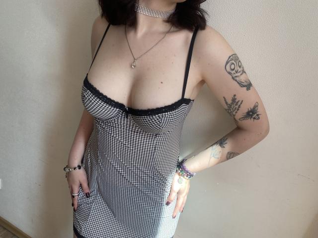 Khori wearing a black and white checkered dress with black lace, showing tits, has tattoos on arms, wearing bracelets, choker, necklace, and ring, standing against a plain wall.