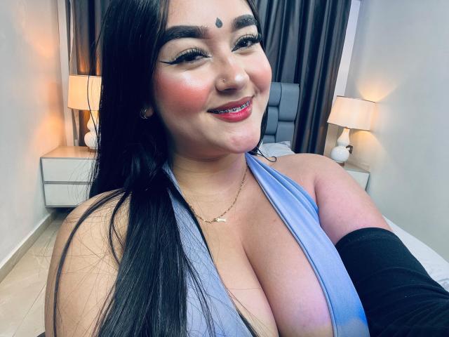 AnaShalimarr, con cabello negro largo, un top azul de tirantes que muestra un busto grande, sonriendo, con lápiz labial rojo, una manga negra en el brazo, un collar dorado, cortinas marrón oscuro y dos lámparas blancas en el fondo..