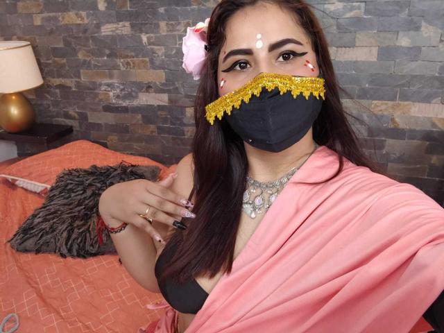 Dalia Dubey, con cabello castaño, usando una máscara negra con detalles amarillos, un sari rosa, un sujetador negro y un collar brillante, mostrando sus senos, y una flor rosa en su cabello..