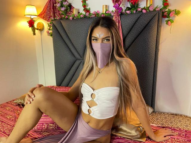 DalilaAliX met lang blond haar, een witte top zonder bandjes met uitsparingen, een paars masker, een paarse rok, zittend op een bed met roze beddengoed met een patroon, kijkend naar de camera, een bloemenhoofddeksel..