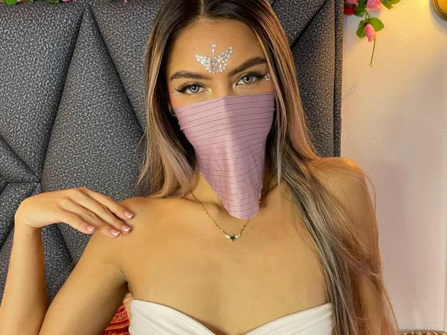 DalilaAliX with long brown hair, wearing a pink mask covering her nose and mouth, white strapless top, and gold necklace, has white butterfly makeup on her forehead.