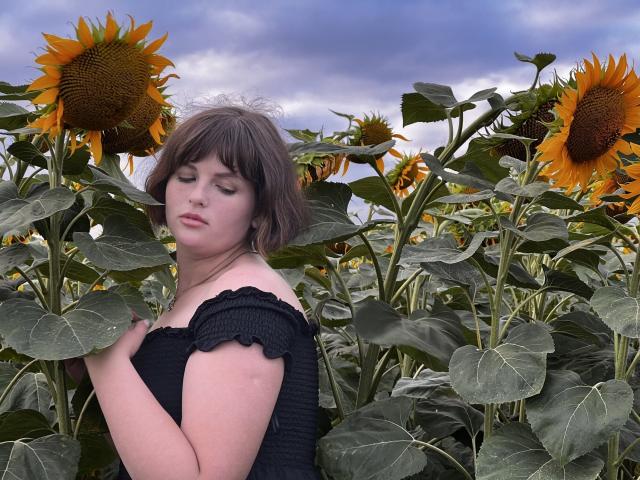 StaceyLex, con cabello castaño corto, ojos cerrados, vistiendo una blusa negra con un hombro descubierto, está de pie en un campo de girasoles, rodeada de plantas verdes altas y girasoles amarillos brillantes..