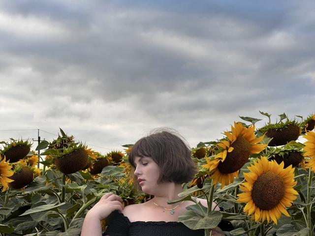 StaceyLex met kort bruin haar, een zwarte top met een schouder, die een zonnebloem vasthoudt, staand in een veld met zonnebloemen, bewolkte lucht, gele bloemblaadjes, groene bladeren, een subtiele ketting, een zachte uitdrukking, een vriendelijke blik..