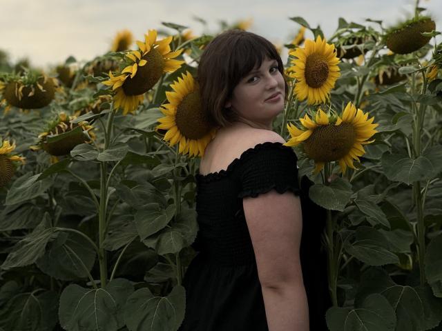 StaceyLex with short brown hair, black off-shoulder dress, standing in sunflower field, looking over shoulder, yellow sunflowers in foreground, green leaves, soft evening light.