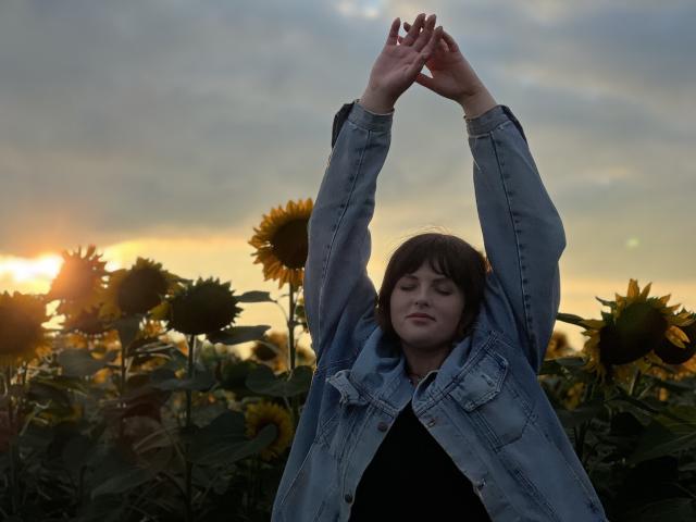 StaceyLex met armen omhoog, een denimjas aan, staand in een zonnebloemveld bij zonsondergang, met gesloten ogen, en een serene uitdrukking. Het zonlicht schijnt achter haar..