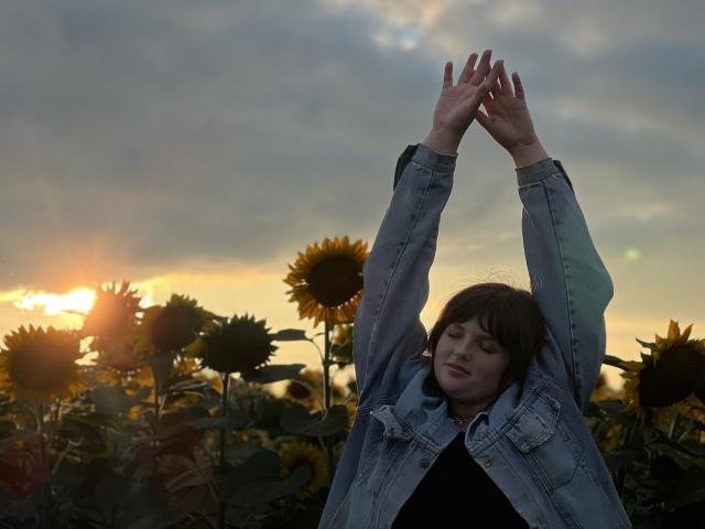 StaceyLex with arms raised, wearing a denim jacket, standing in a sunflower field at sunset, eyes closed, hands touching, short brown hair, calm expression.